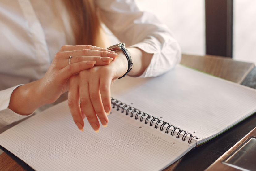Time management - Image of young Caucasian women sitting at a desk while looking at her wrist watch with her left arm resting on a notebook