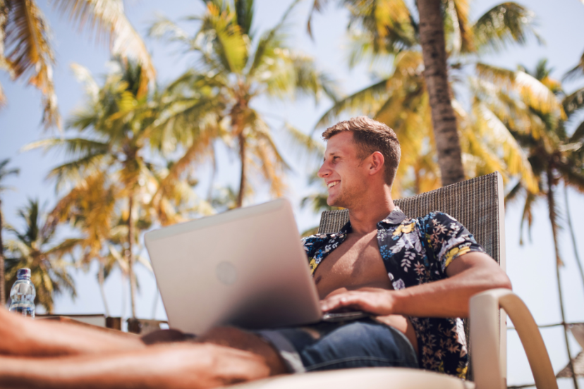 Digital nomad lifestyle - Image of young man in a pair of short with a tropical pattern shirt exposing his chest while reclined on a lounge chair on a tropical beach with palms tree and working on his laptop