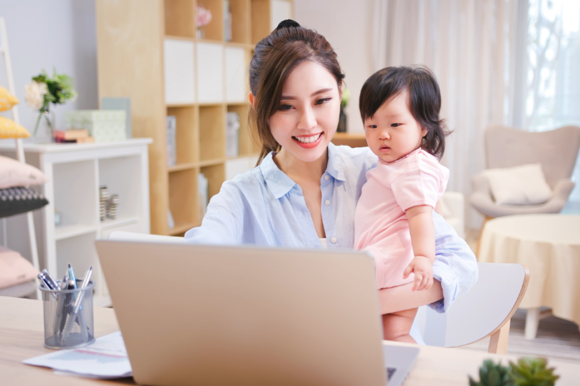 Remote Work: Young Asian Woman holding her child in her left arm while working on her laptop from her home office