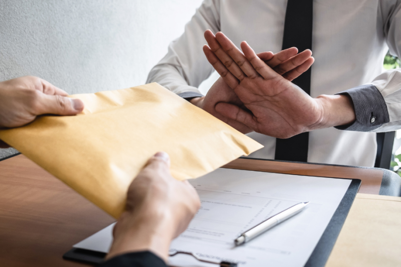 Work-from-home scams - Image of two men facing each at a desk with one man crossing his hands in response to the other man offering something in a business sized envelope