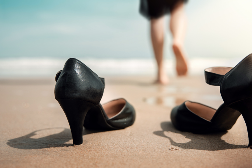 Work-Life Balance - Young woman wearing a black dress who decides to take her black high healed off before walking into the ocean barefoot